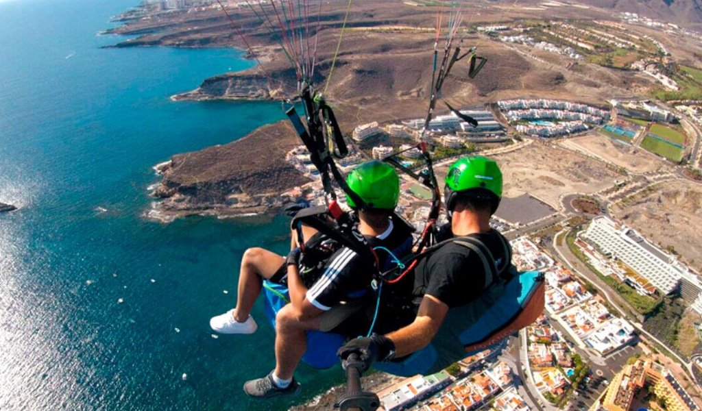 Personas haciendo parapente sobre la costa de Tenerife con vista aérea del mar y edificios.