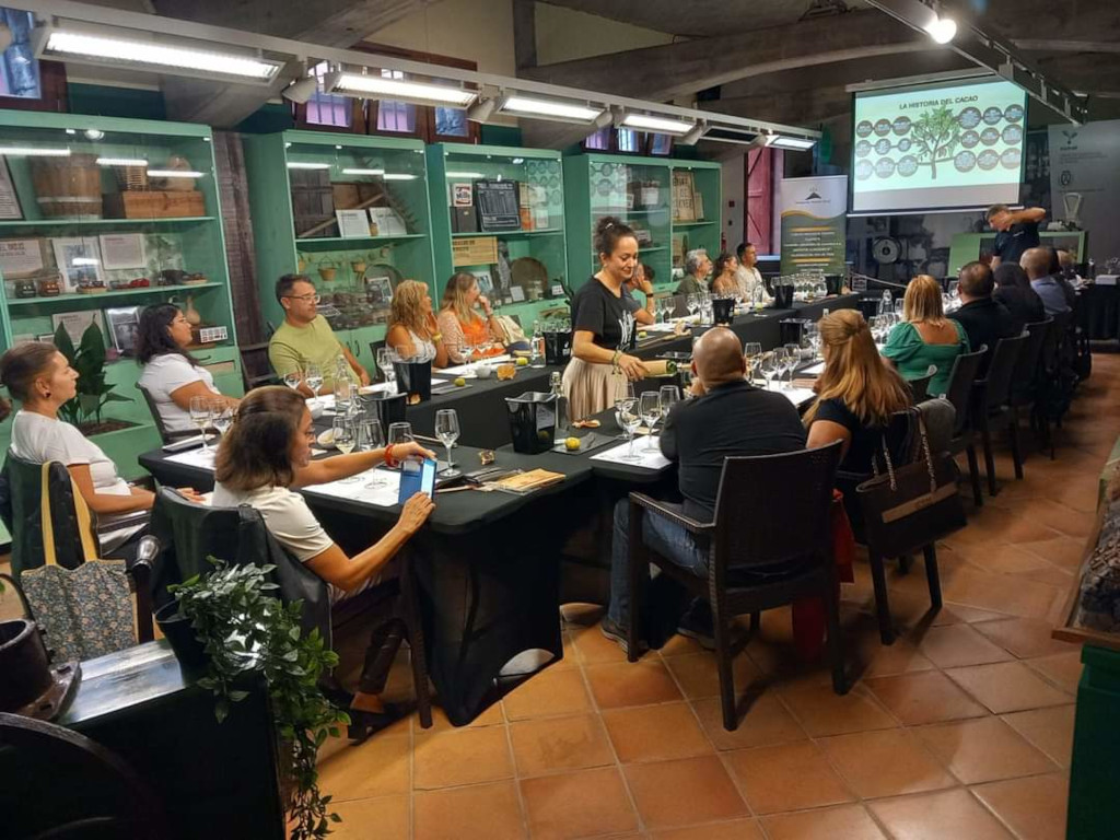 Personas en una cata de vinos en una sala decorada en Tenerife. Texto visible: La Anatomía del Cacao.
