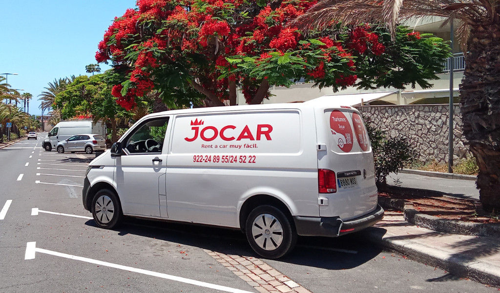 Furgoneta de alquiler Jocar en Santa Cruz de Tenerife estacionada junto a árbol con flores rojas.