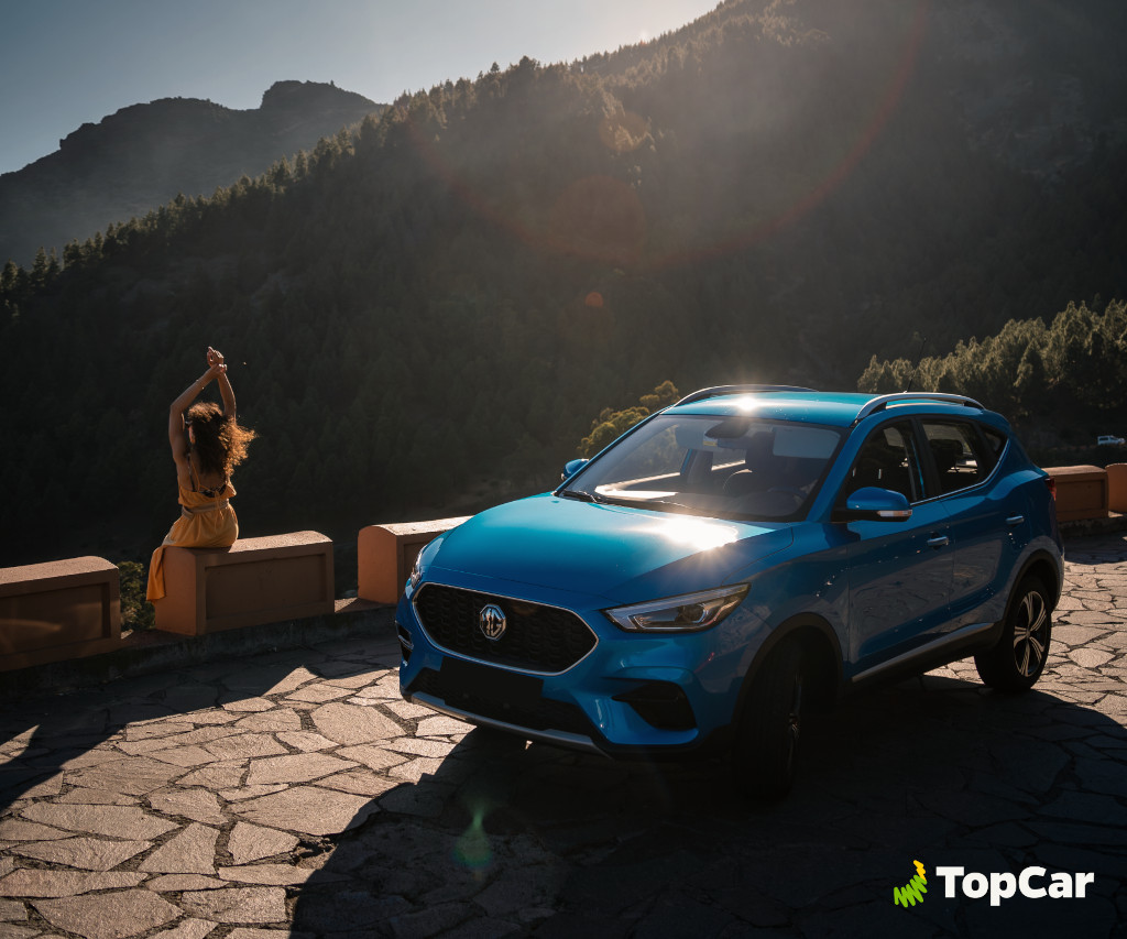 Coche azul frente a montañas en Tenerife, mujer sentada en muro bajo el sol.