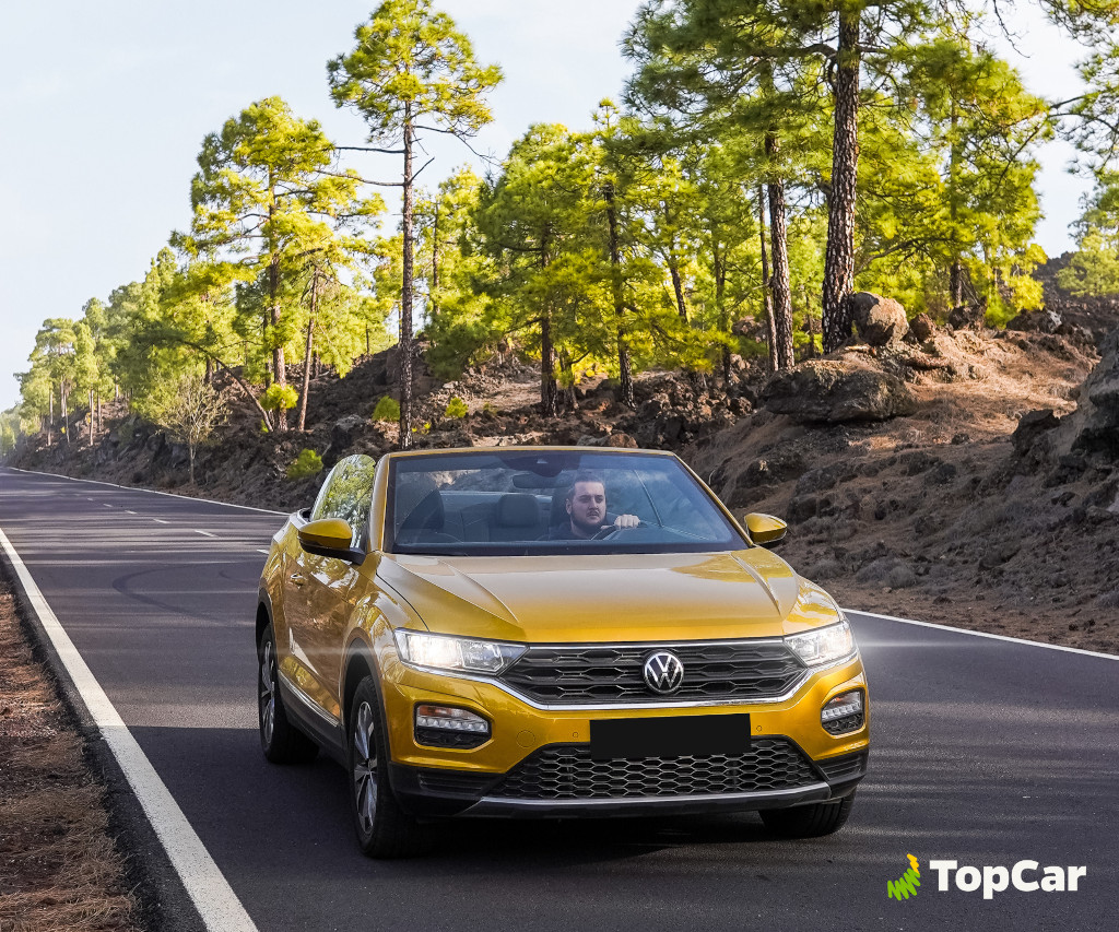 Coche amarillo en carretera rodeada de pinos en Tenerife. Logo de TopCar.