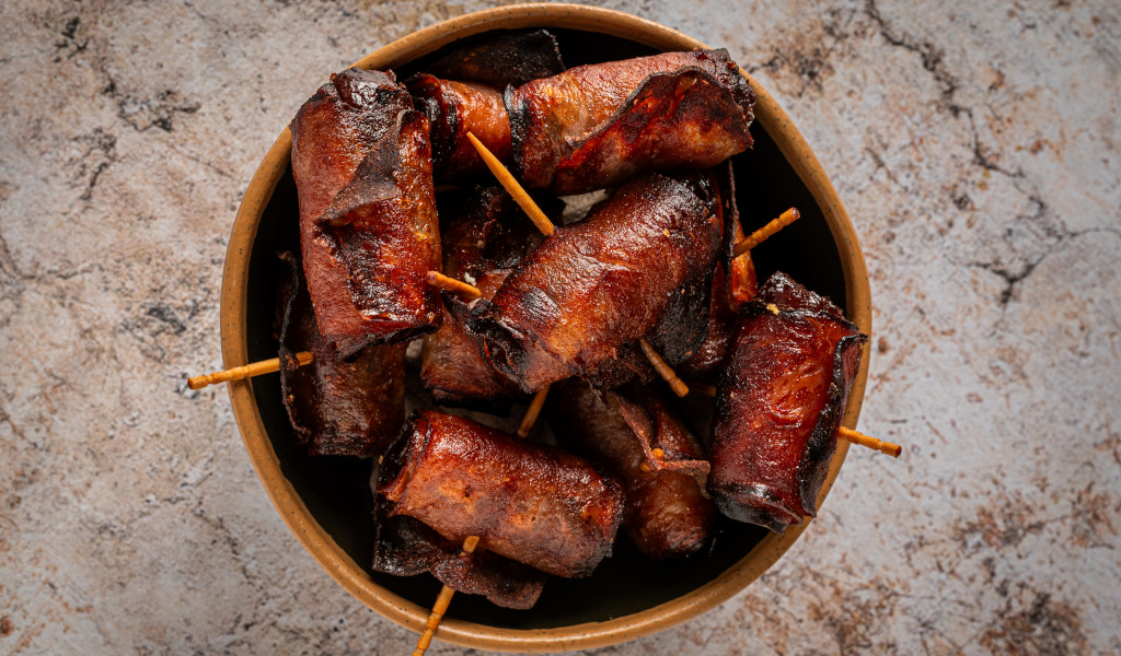 Salchichas envueltas en tocino con palillos sobre fondo de piedra.