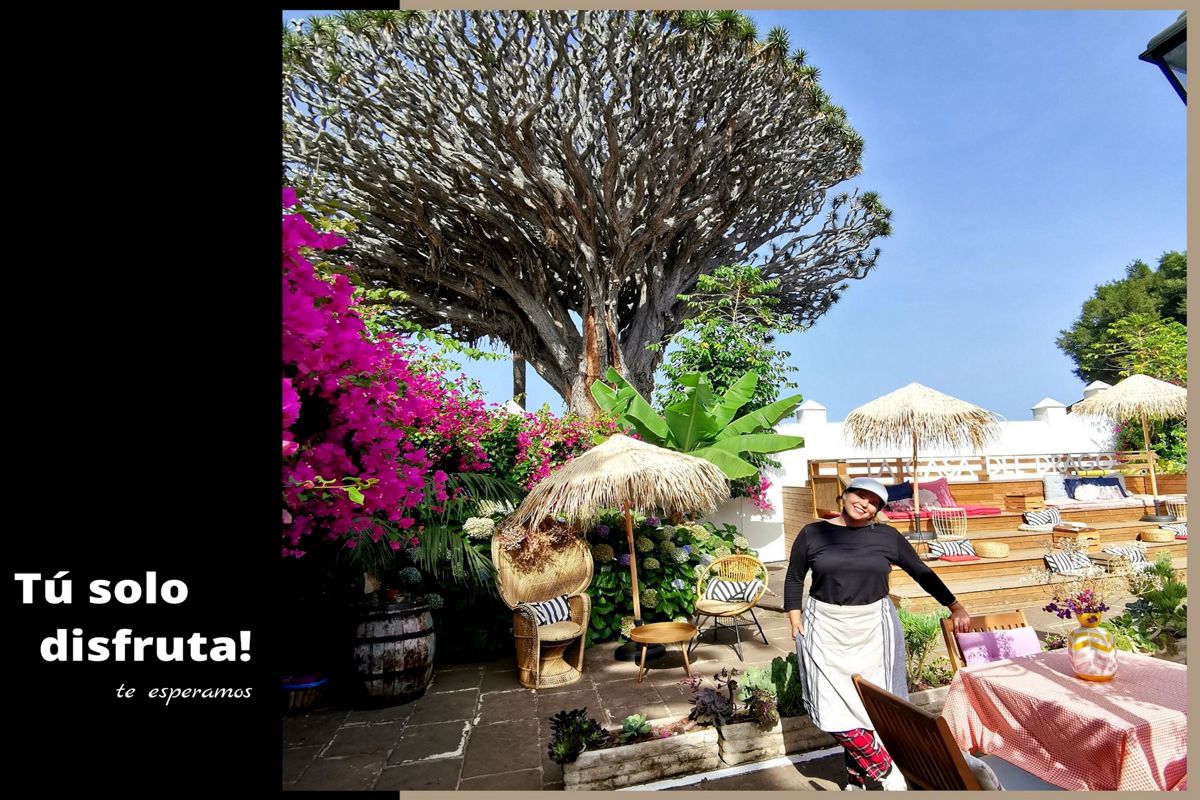 Patio con vegetación, sombrillas de paja y una persona en Tenerife. Texto: Tú solo disfruta! te esperamos.