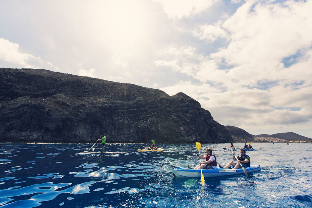 Personas practicando kayak y paddle surf en la costa de Tenerife con montañas al fondo.