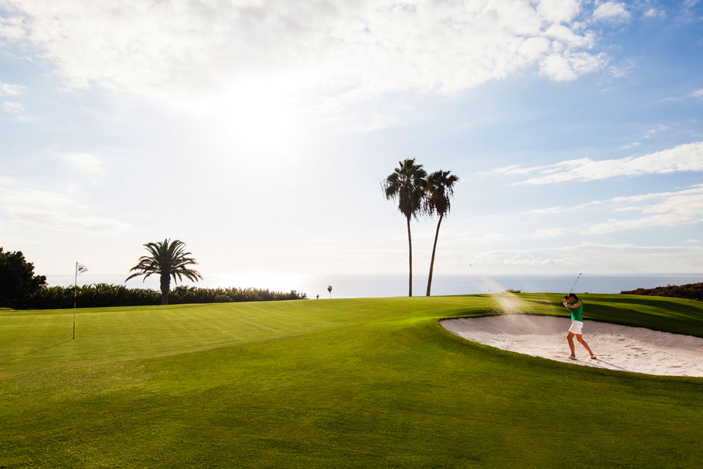 Persona jugando golf en un campo junto al mar en Tenerife con palmeras y cielo despejado.