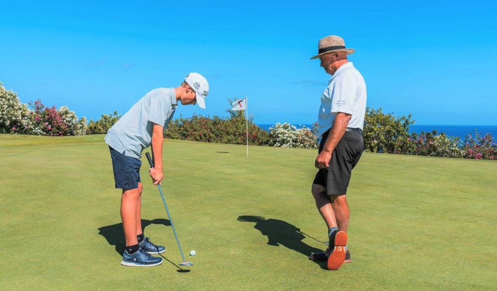 Niño practicando golf con adulto en campo, Tenerife de fondo.