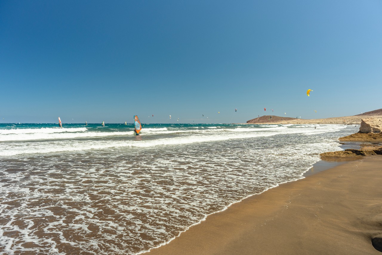 Surfistas en la playa de El Médano, Tenerife, con parapentes en el cielo.