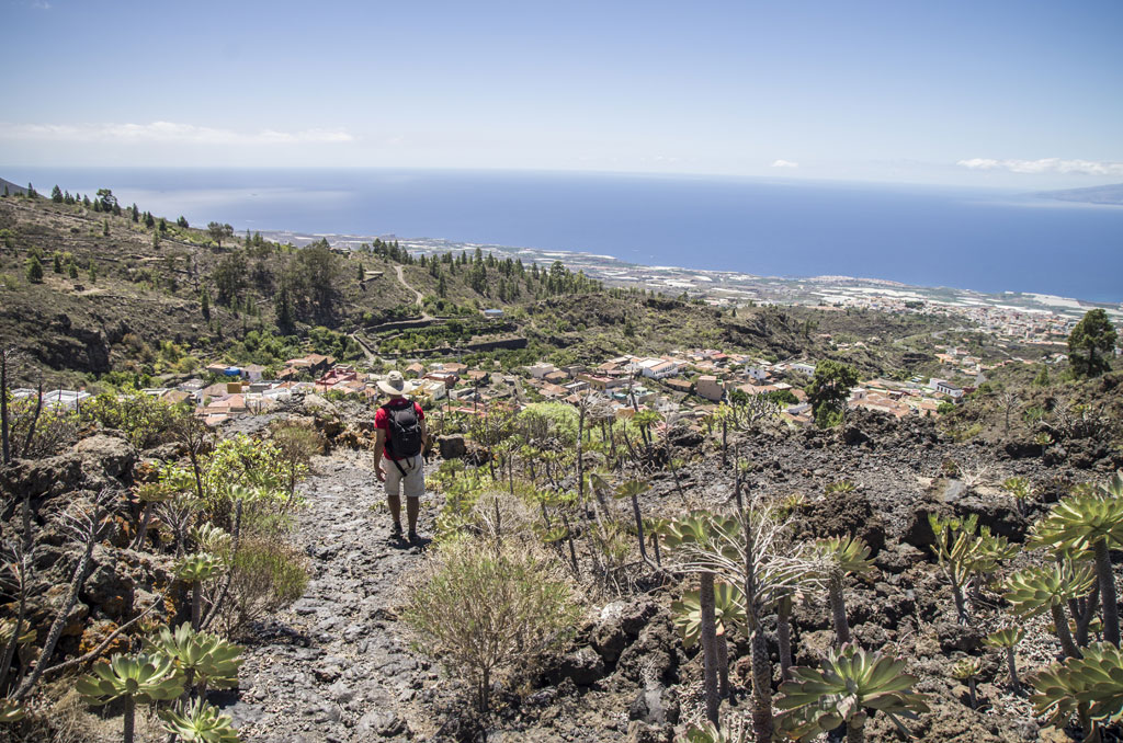 Senderista recorriendo un sendero rocosa con vistas a un pueblo y el mar en Tenerife.