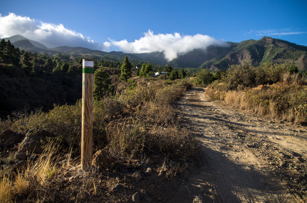 Sendero de tierra con paisaje montañoso en Tenerife, poste de señalización a la izquierda.