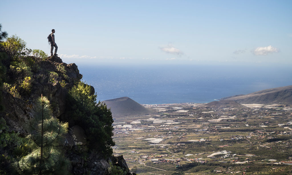 Persona contemplando el paisaje desde una cima en Tenerife, con vista al océano y campos.