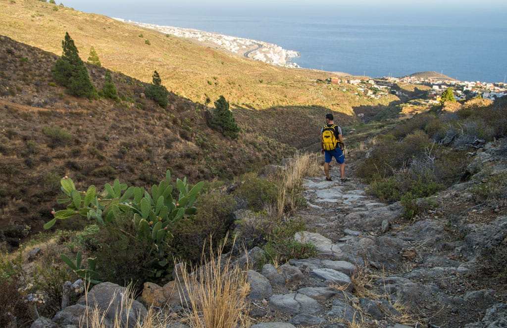 Persona caminando en sendero hacia el mar en el paisaje montañoso de Tenerife.