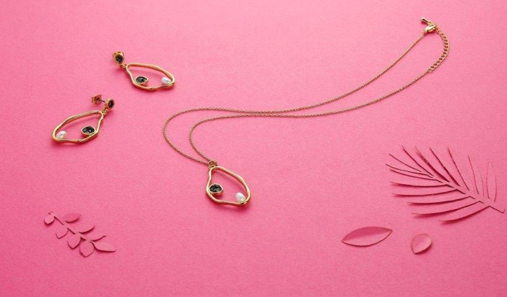 Collar y pendientes de oro con perlas sobre fondo rosa y decoraciones de hoja.