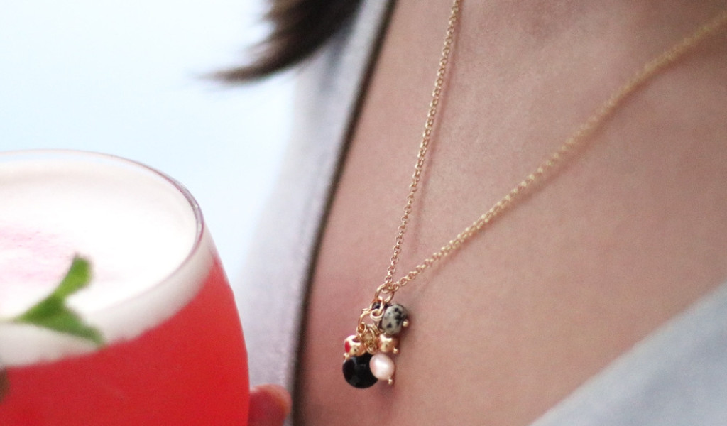 Collar con colgantes y bebida rosada en vaso junto a una persona.