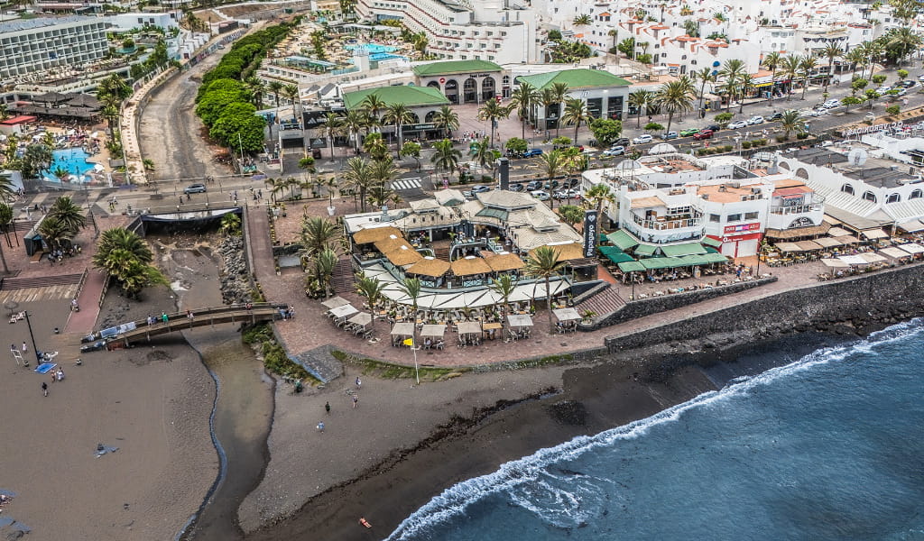 Vista aérea de playa y zona urbana en Tenerife con edificios y restaurantes cercanos.