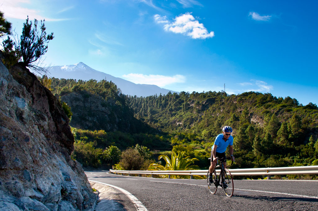 Ciclista en carretera rodeada de montañas y vegetación, con vista al Teide en Tenerife.