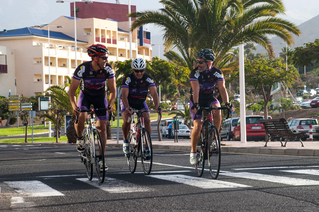 Ciclistas cruzan la calle en Las Américas, Tenerife, con palmeras de fondo.