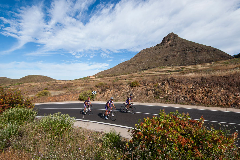 Ciclistas en bicicleta por carretera en Tenerife, paisaje montañoso al fondo.