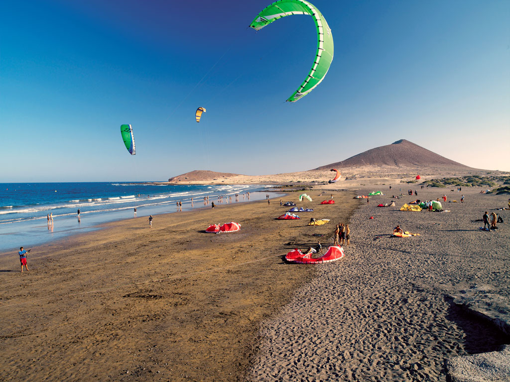 Personas practicando kitesurf en la playa de El Médano, Tenerife, con montañas al fondo.