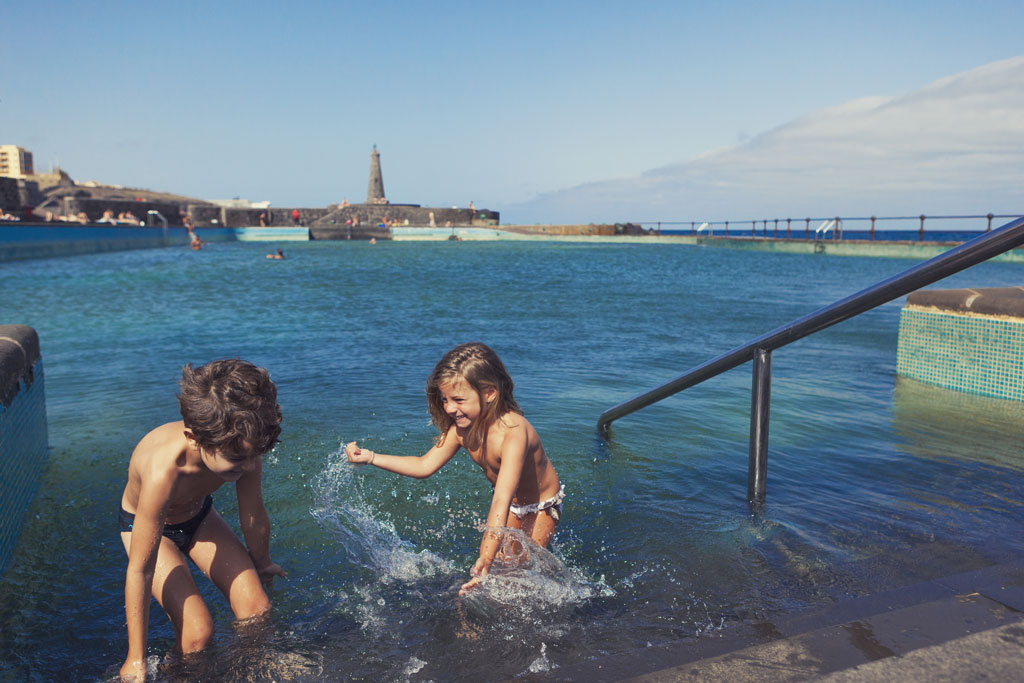 Niños jugando en una piscina natural de Bajamar, Tenerife, con un faro al fondo.
