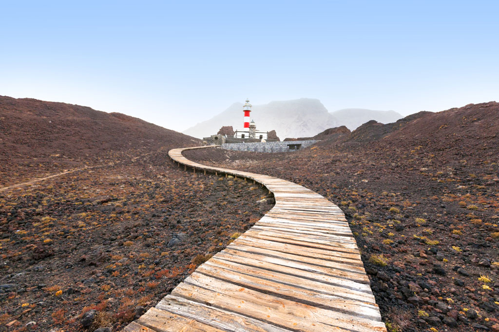 Pasarela de madera hacia el faro de Punta de Teno, Tenerife, con montañas al fondo.