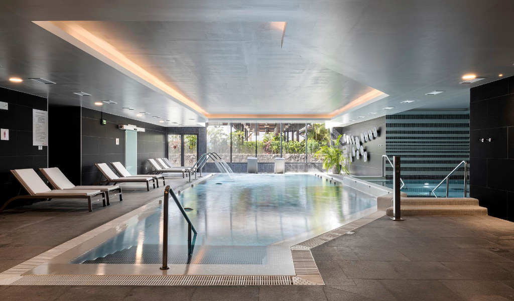 Piscina interior en Tenerife con tumbonas y plantas junto a las ventanas.