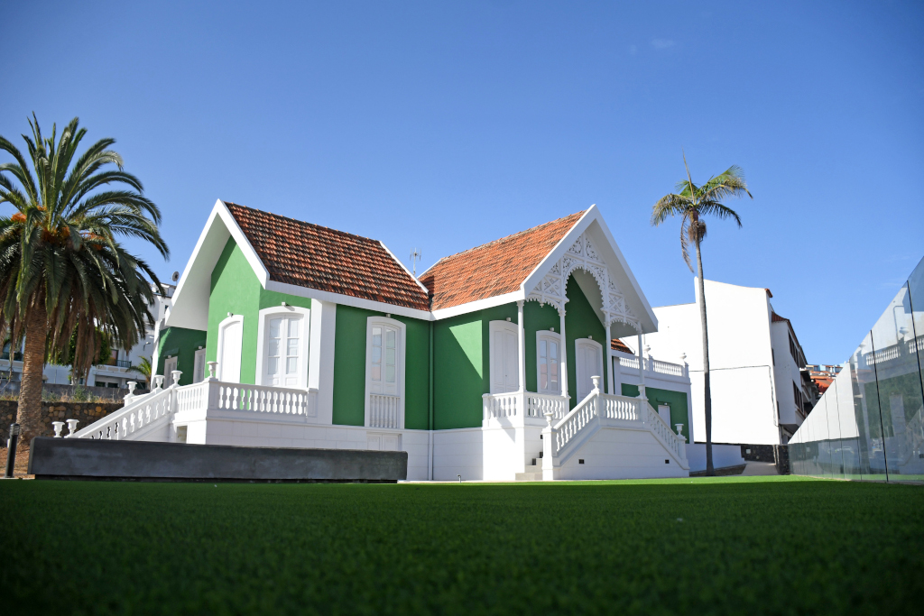 Casa antigua verde y blanca con techo rojo en Tenerife, rodeada de palmeras y césped.